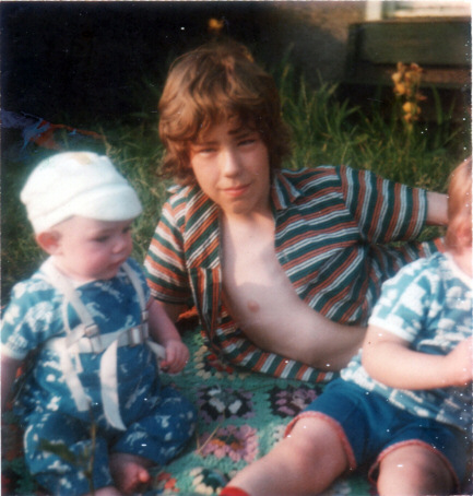 Picture of a teenage boy lying on the grass with his shirt undone, and with two small boys in the picture