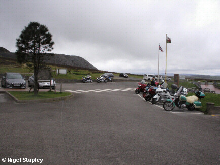 Photo across a small car park towards the spoil heaps of an old slate quarry. Includes motorbike with a sheepskin seat-cover